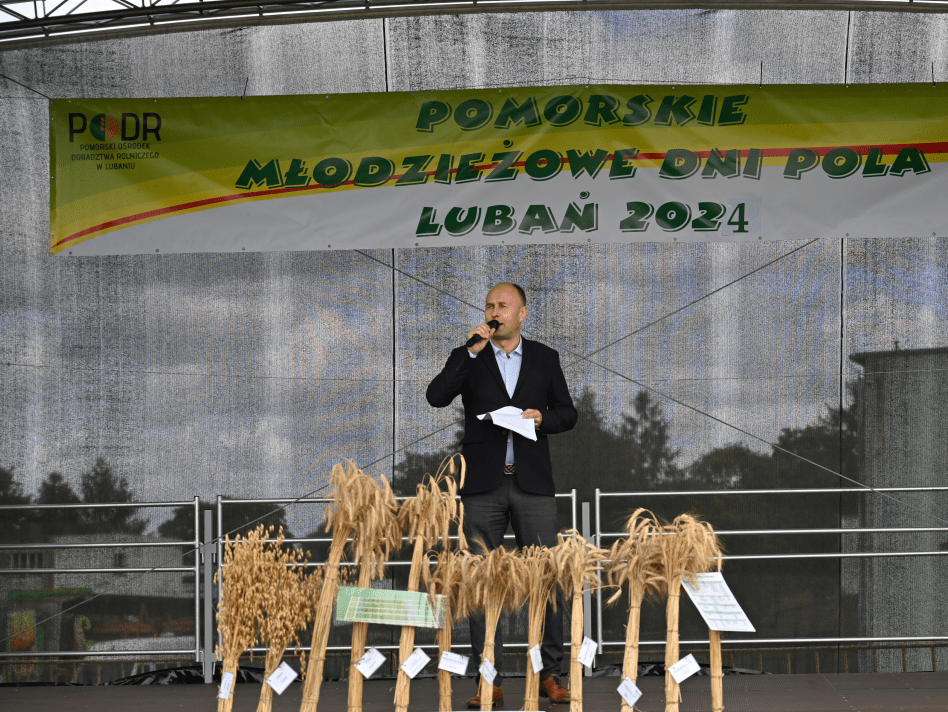 Na zdjęciu widzimy Dyrektora PODR w Lubaniu, Aleksandra Macha, przemawiającego na scenie podczas oficjalnego otwarcia IV Pomorskiego Młodzieżowego Dnia Pola. W tle znajduje się transparent z nazwą wydarzenia, a na pierwszym planie eksponowane są różne odmiany zbóż, ilustrujące różnorodność lokalnych upraw.