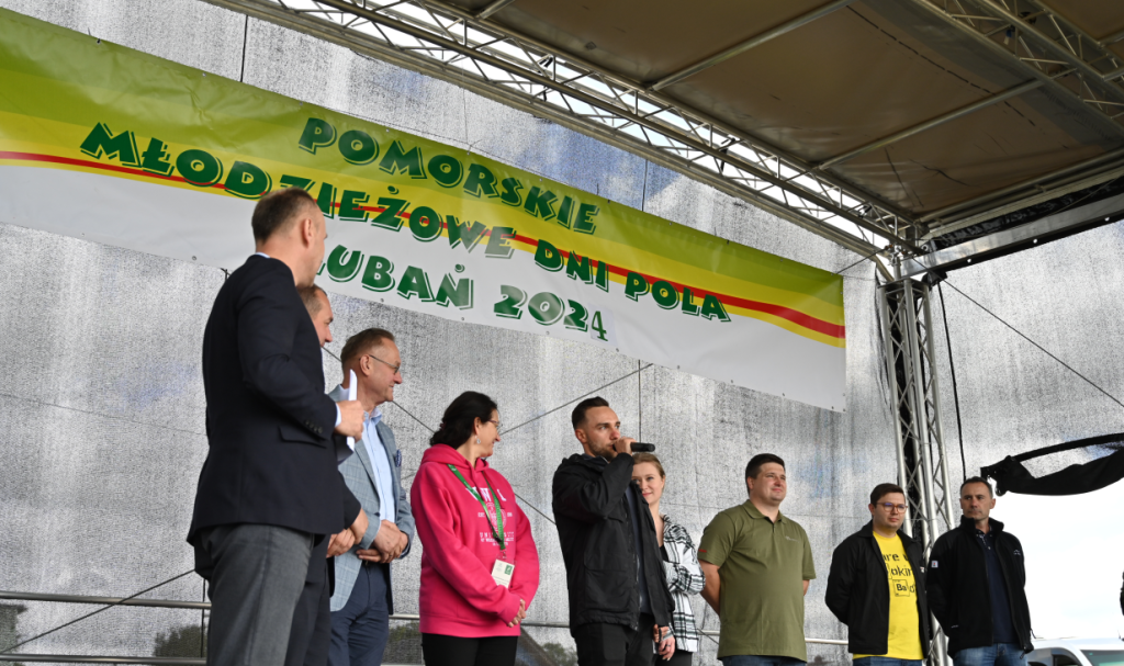 Na scenie stoi grupa organizatorów i specjalistów biorących udział w wydarzeniu. Wśród nich można zauważyć ekspertów z różnych instytucji oraz firm, którzy wspierali organizację i prezentacje podczas Młodzieżowego Dnia Pola. Transparent w tle informuje o nazwie i roku wydarzenia.