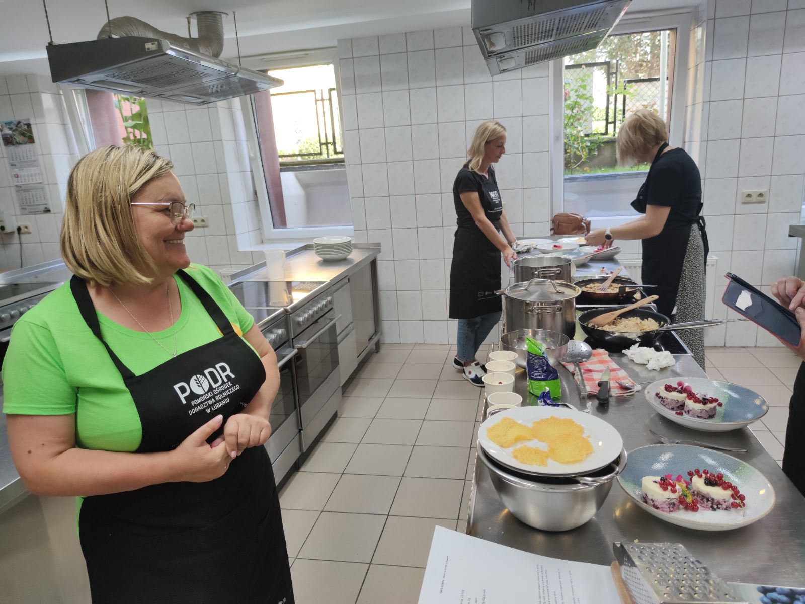 Grupa kobiet ubranych w fartuchy z logo PODR pracuje nad przygotowaniem różnych dań w profesjonalnie wyposażonej kuchni. Jedna z uczestniczek z uśmiechem obserwuje postępy innych. Na stole widać gotowe potrawy, w tym desery z owocami.